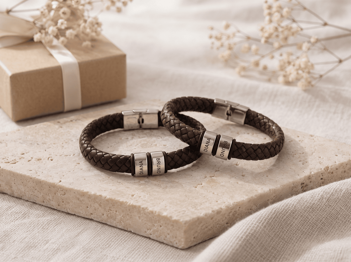 Two dark brown braided leather bracelets with silver-toned metal accents engraved with names, displayed on a beige stone slab with a kraft gift box and dried flowers in the background.