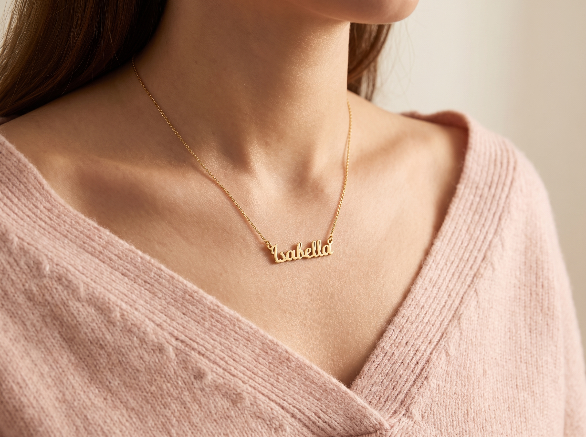 A close-up of a model wearing a delicate gold nameplate necklace that spells 'Isabella' on a soft pink knit sweater.