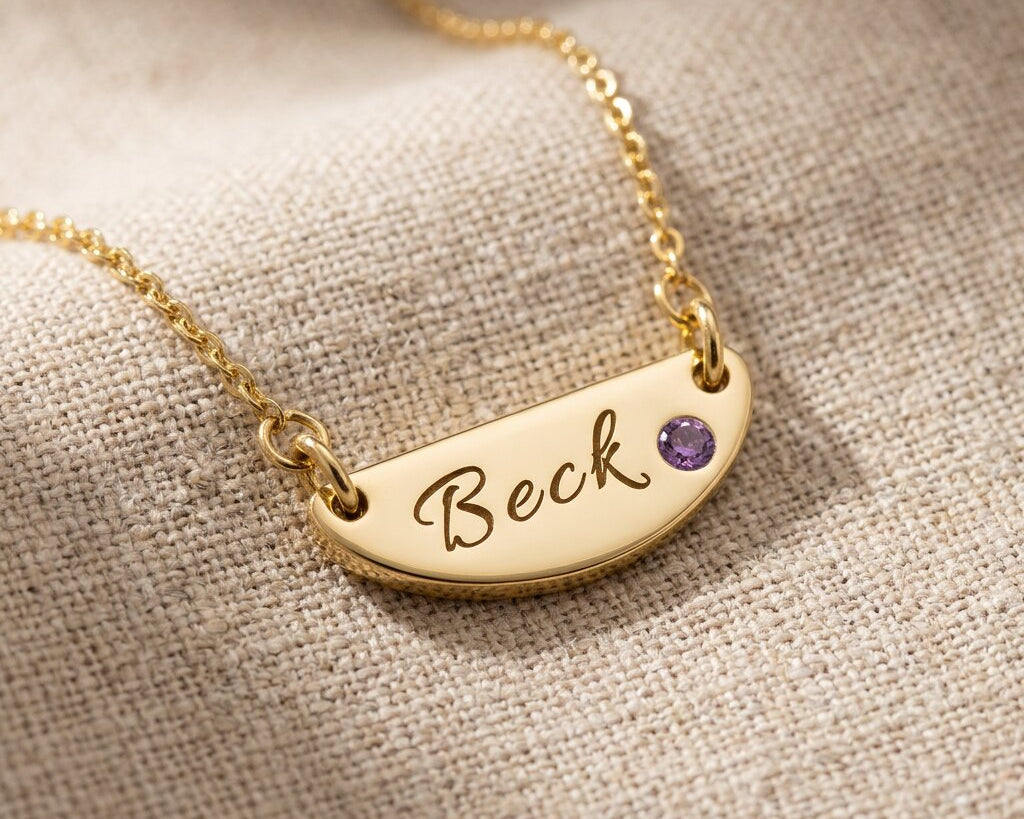 Gold Beck name necklace with engraved bar pendant and birthstone displayed on neutral fabric background
